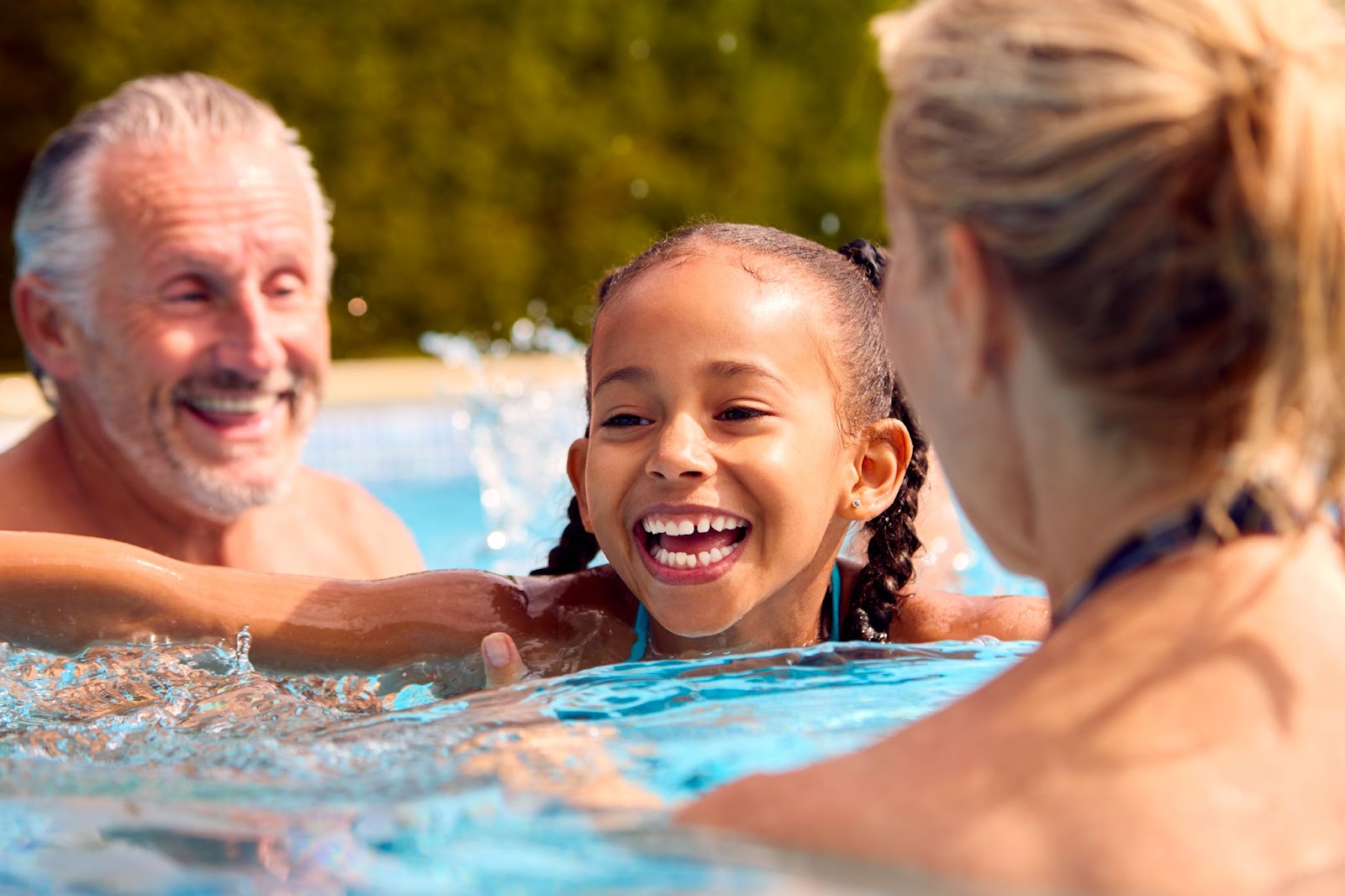 A family enjoying time in a pool, featuring a little girl splashing and an older man relaxing nearby.