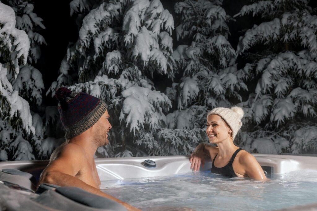 A man and woman relax in a hot tub surrounded by snow-covered trees enjoying a serene winter landscape
