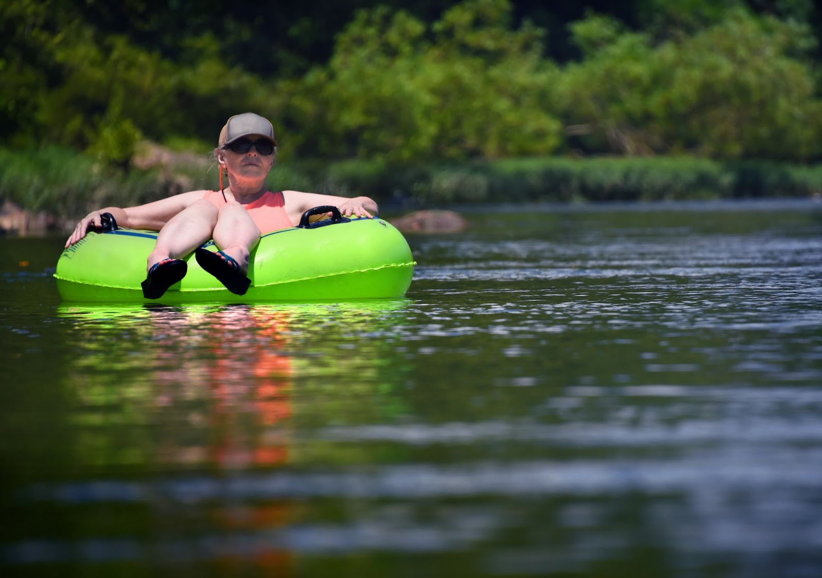 A person wearing sunglasses and a hat floating peacefully down a calm river in a bright green inflatable inner tube.