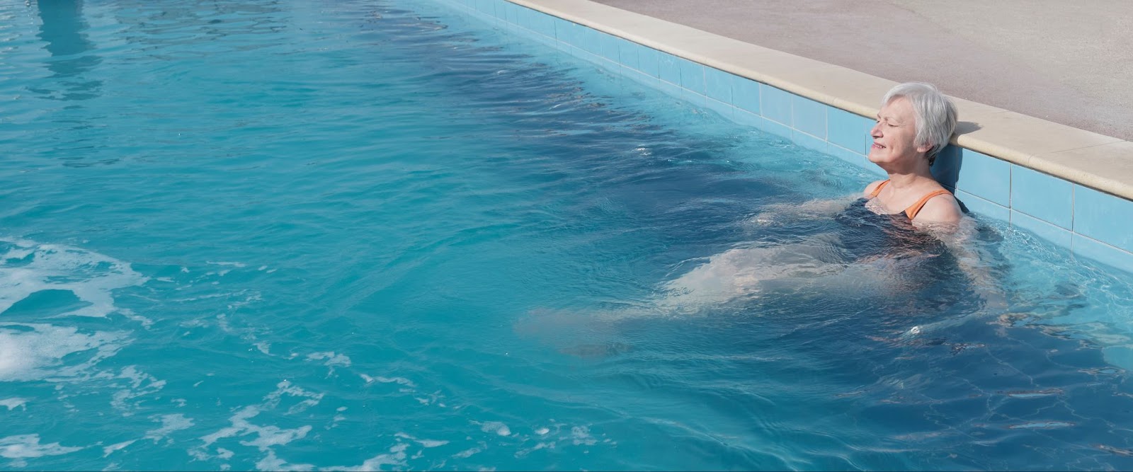 An elderly woman with short grey hair relaxing in a bright blue swimming pool with her eyes closed and back against the edge.